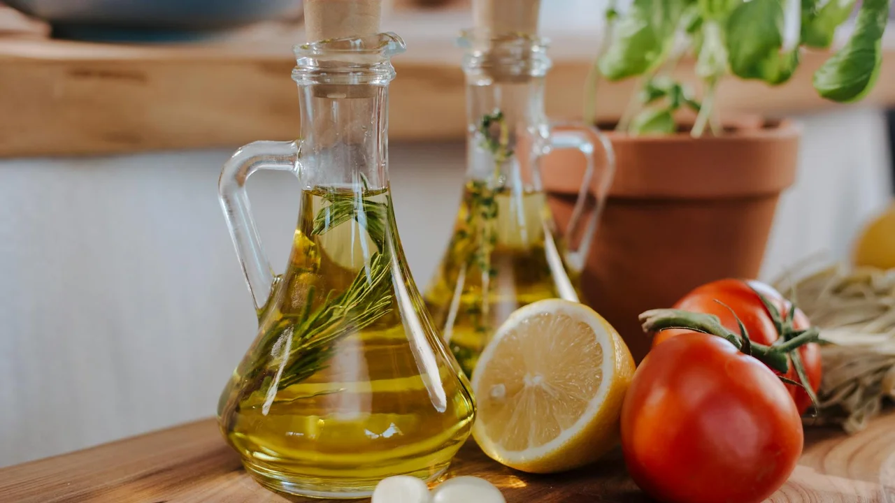 Olive oil in glass bottles with a halved lemon, tomatoes, and fresh herbs on a wooden kitchen counter.