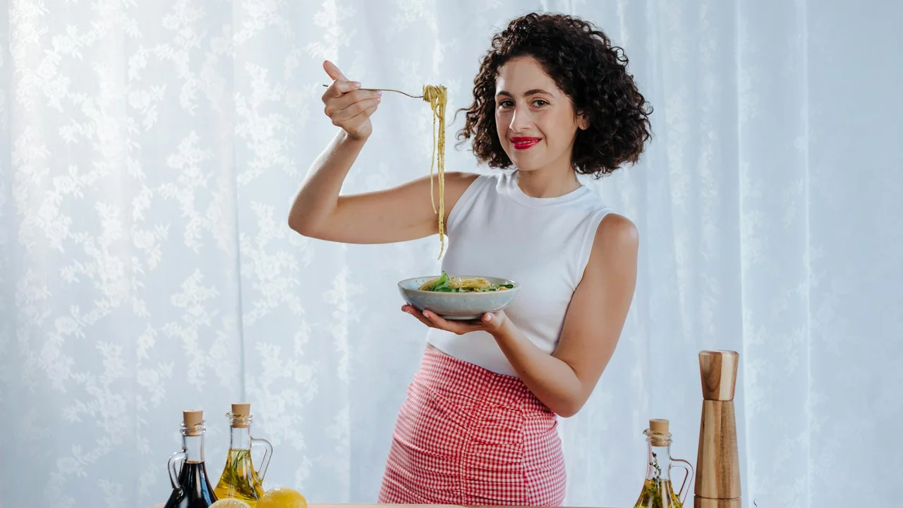 A woman in a white sleeveless top and red checkered skirt stands in a bright kitchen, lifting noodles with chopsticks from a bowl; olive oil bottles and kitchen tools are on the counter behind her.