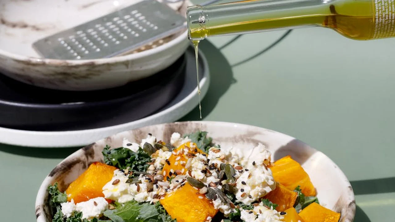 A stream of olive oil being poured from a bottle toward a pan, with a plate of cubed squash and crumbled cheese in the foreground.