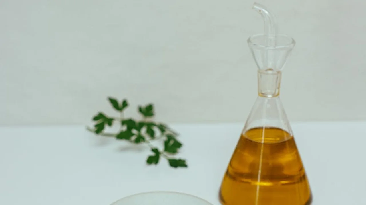 A glass bottle filled with golden olive oil and a pour spout sits on a white surface, with a small sprig of herbs in the background.