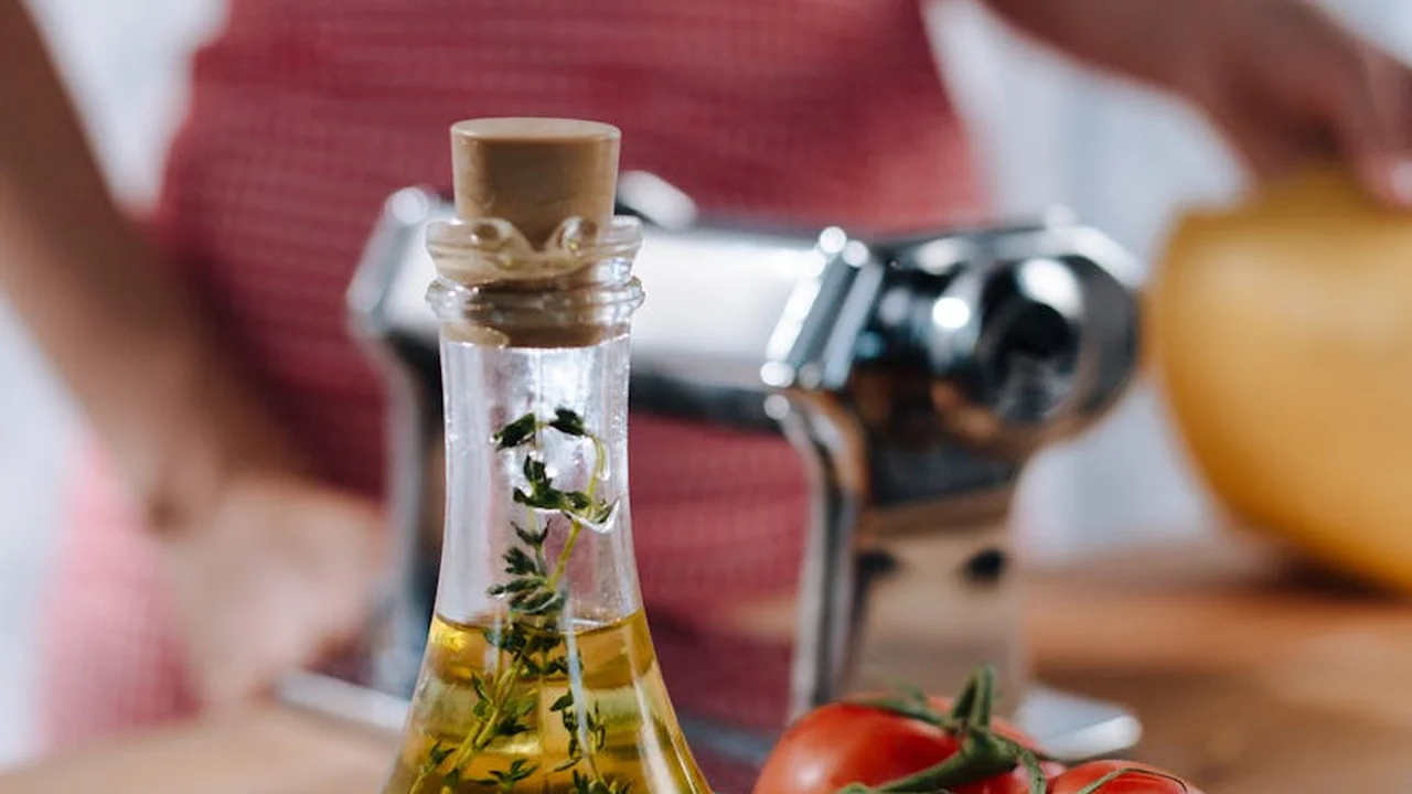 Close-up of a glass bottle of olive oil with a cork stopper on a kitchen counter, with a blurred background of a metal appliance and other ingredients.