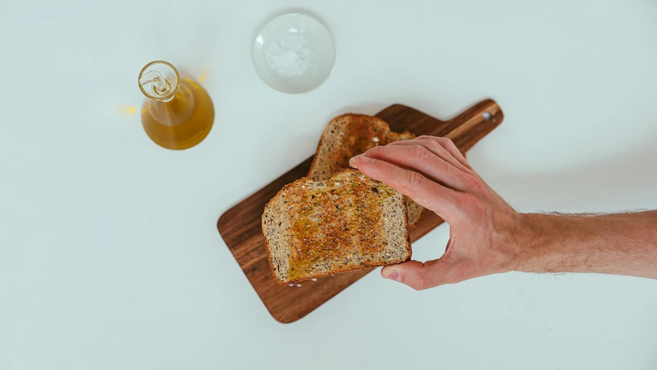 A hand reaches for a slice of bread on a wooden cutting board with a small bottle of olive oil and a glass nearby on a light surface.