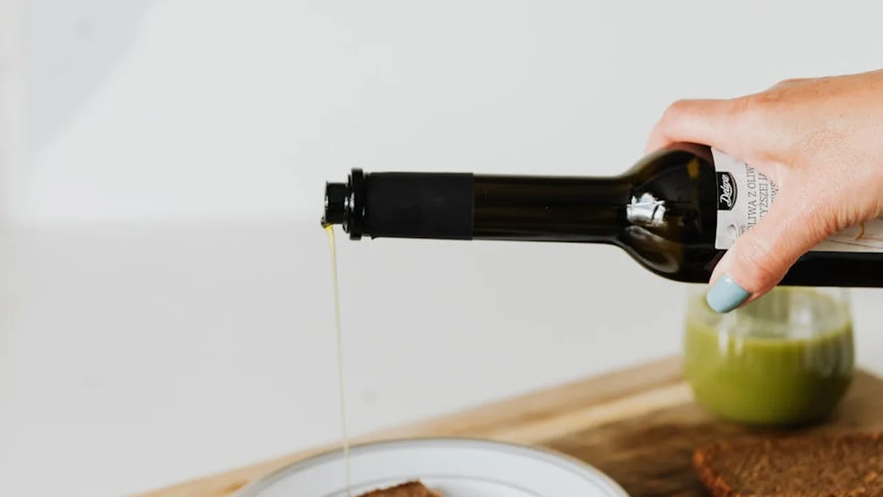 A hand pours olive oil from a dark bottle into a small dish on a wooden countertop, with a jar of green sauce in the background.