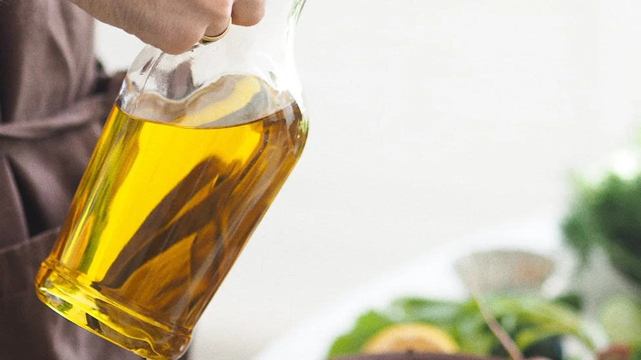 Close-up of a hand pouring golden olive oil from a glass bottle in a bright kitchen, with blurred vegetables in the background.