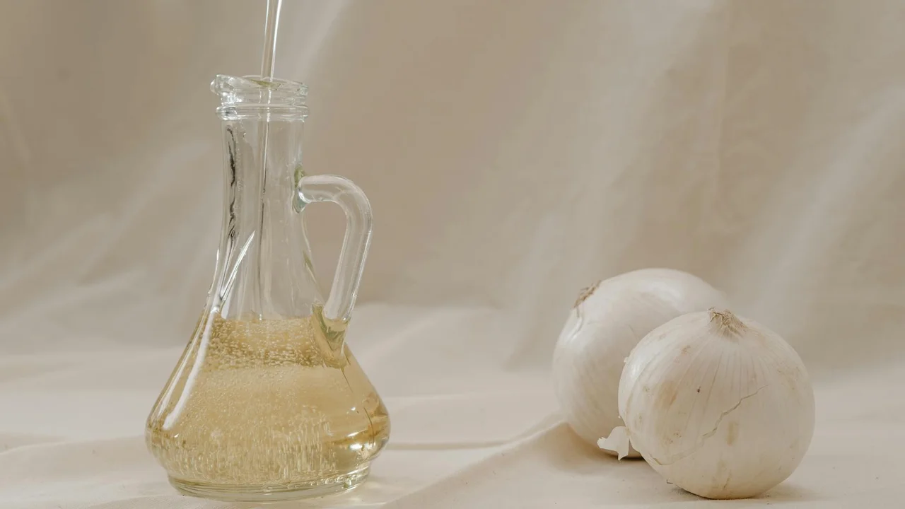 Clear glass bottle of olive oil with a spout and handle on a light beige surface, accompanied by two white onions.
