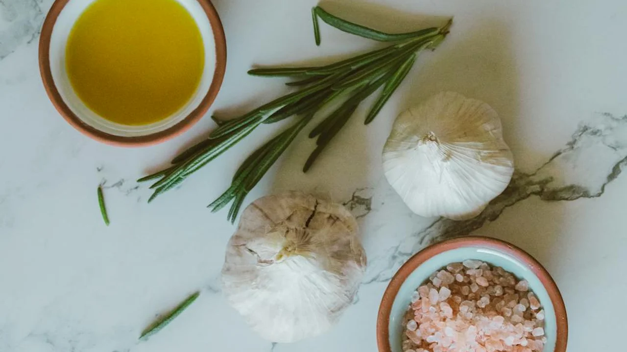A small bowl of olive oil, sprigs of rosemary, two garlic bulbs, and a dish of pink salt on a marble countertop