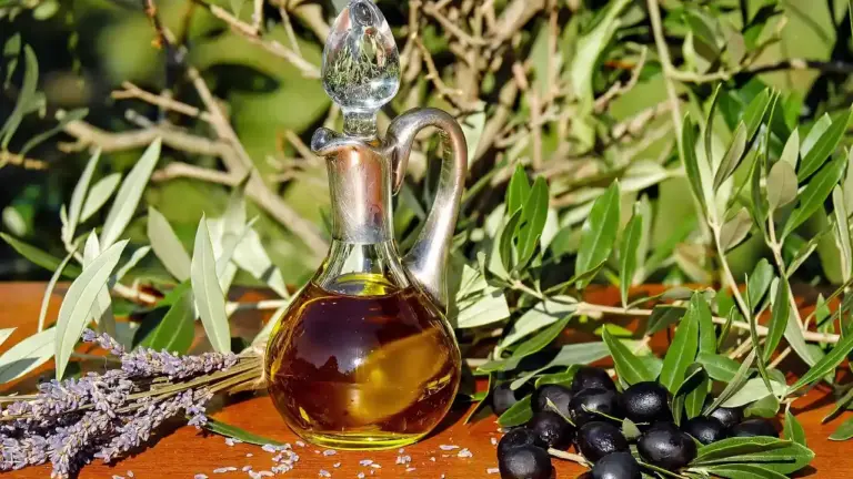 Glass bottle of olive oil with a decorative stopper on a wooden surface, surrounded by olive branches, green leaves, olives, and sprigs of lavender