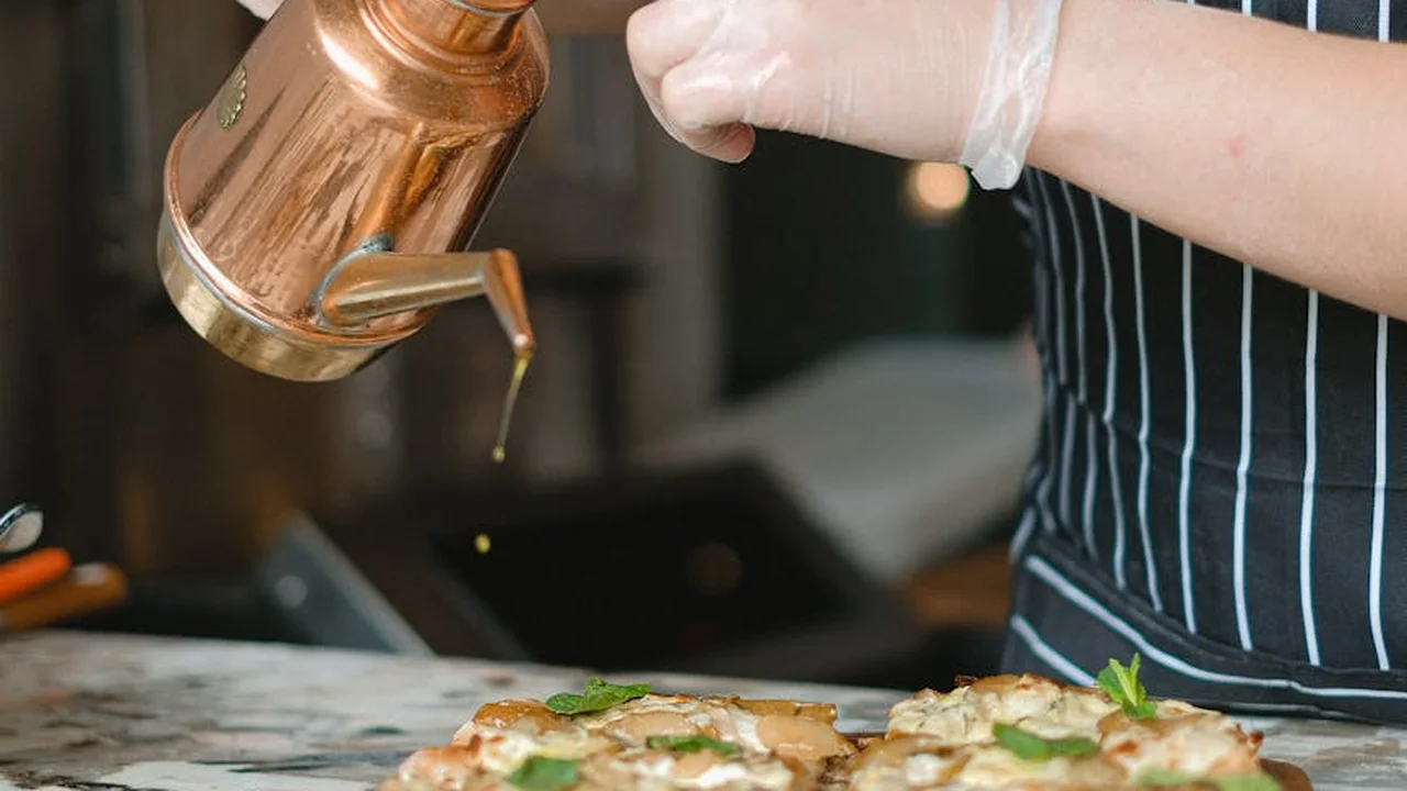 Chef wearing gloves and a striped apron pours olive oil from a copper pitcher onto herbs arranged on a cutting board.