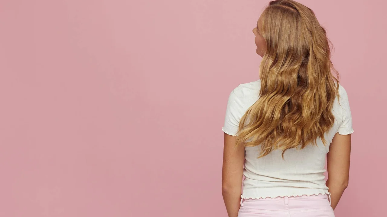 Rear view of a woman with long, wavy blonde hair wearing a white top against a pink background.