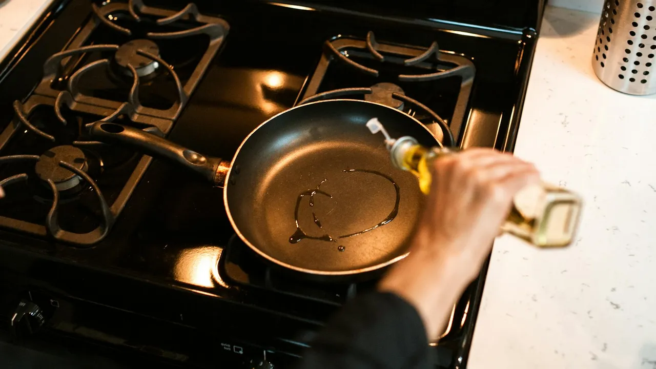 A hand pouring oil into a hot frying pan on a gas stove.