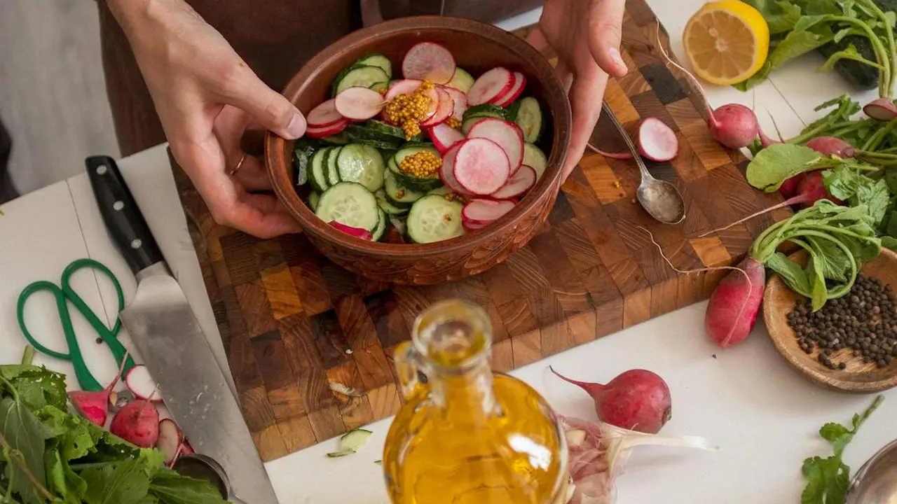 Hands hold a basket of sliced cucumbers and radishes over a wooden cutting board, with a bottle of olive oil and fresh herbs nearby.