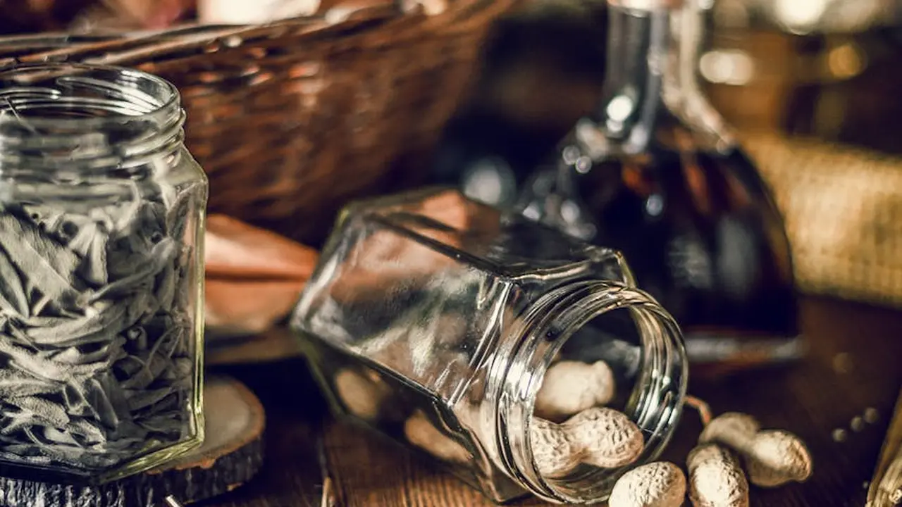 A glass jar tipped over with peanut shells spilling onto a dark wooden surface, with another jar and basket in the background.
