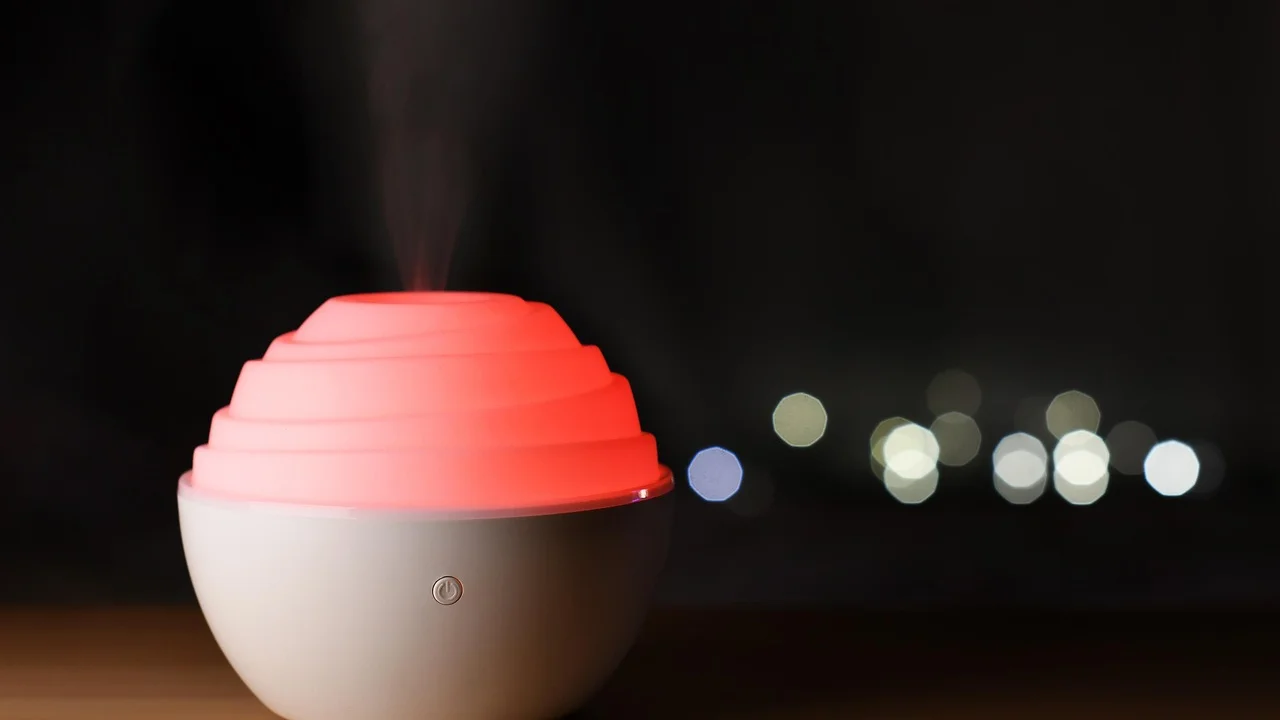 Round white diffuser with a pink ribbed top emitting a gentle mist, set against a dark background with blurred city lights.