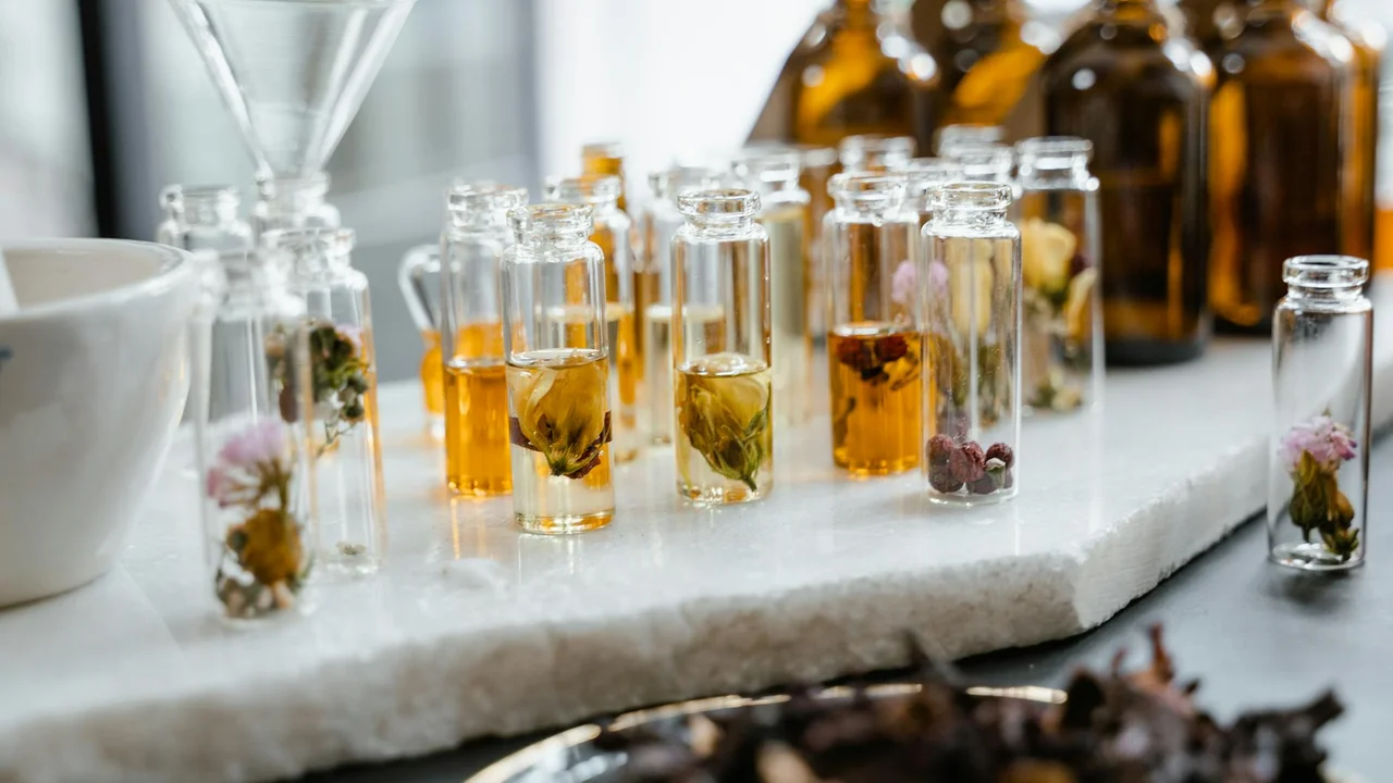 Small glass vials containing pale yellow essential oil with dried botanicals inside, arranged on a white marble surface with larger amber bottles blurred in the background.