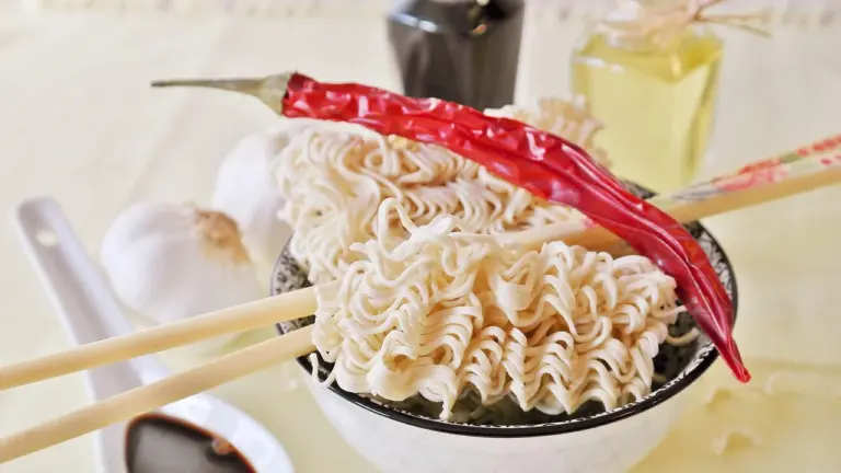 Bowl of ramen noodles with chopsticks resting on the side and a red chili pepper on top.