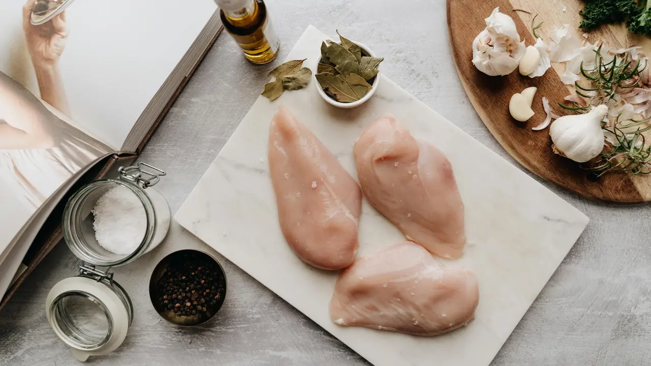 Three raw chicken breasts on a white cutting board with garlic, herbs, and spices nearby, ready for frying.