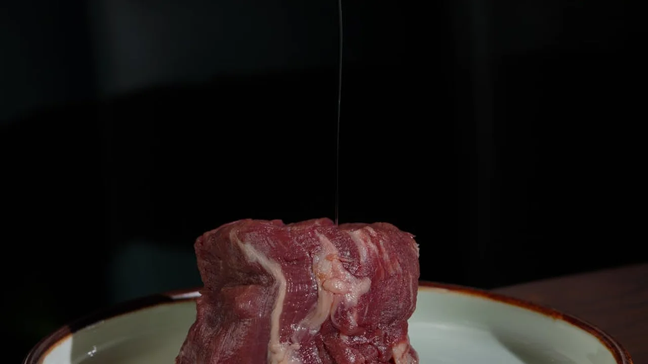 Raw piece of meat on a white plate with a thin stream of oil being poured over it.