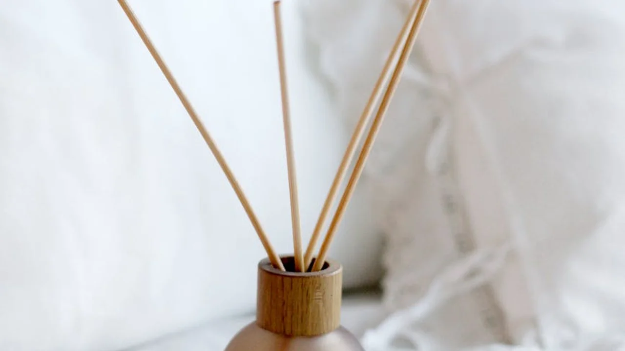 Reed diffuser in a glass bottle with a wooden cap and light-colored reeds against a soft white background.