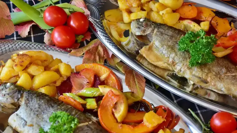 Two plates of roasted fish with a colorful mix of peppers, potatoes, cherry tomatoes, and a sprig of parsley.