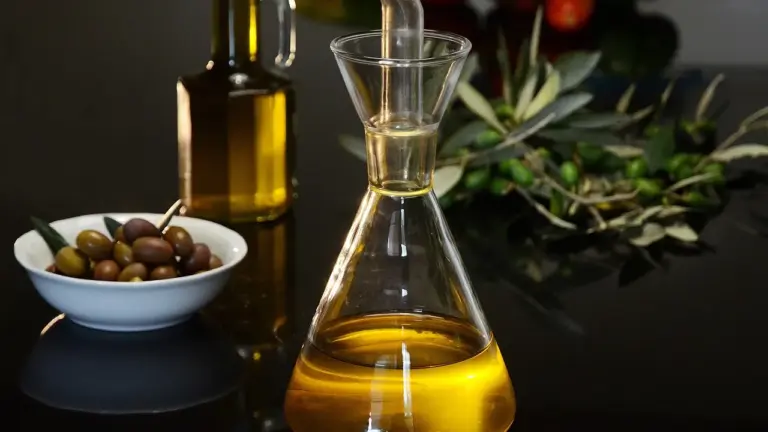 A glass cruet of golden olive oil with a bowl of olives and olive branches in the background, illustrating cooking, dressing, and dipping oils.