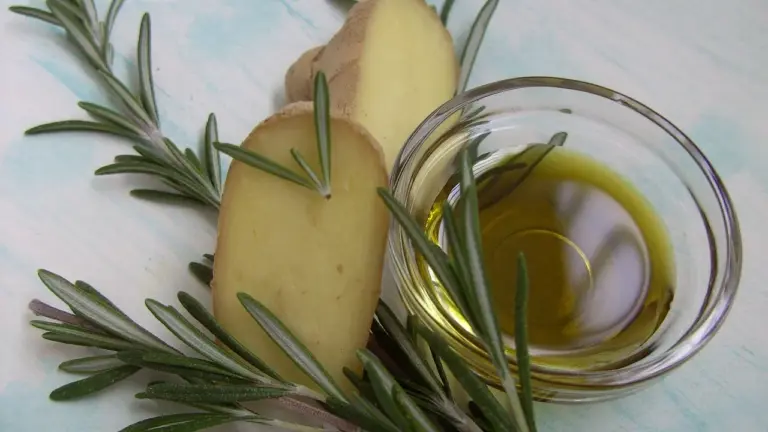 Rosemary sprigs beside a small glass bowl of oil on a light background