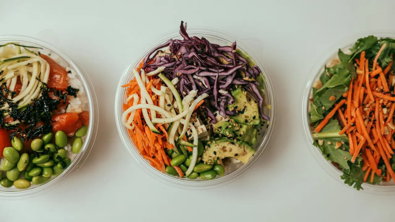 Three clear bowls filled with colorful salad ingredients on a light background, including greens, shredded purple cabbage, carrots, edamame, and other vegetables.