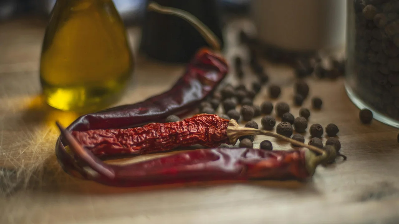 Dried red chili peppers and peppercorns on a wooden surface with a bottle of olive oil in the background.