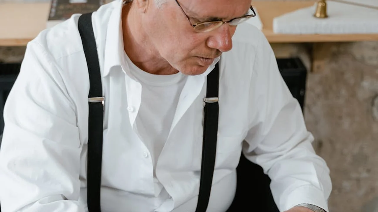 Man in a white shirt with suspenders looking down in a workshop-like setting.