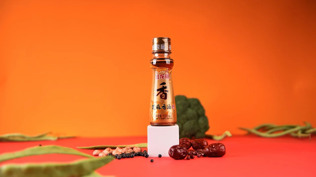 A bottle of sesame oil centered on a bright orange background with spice accents and red dried peppers