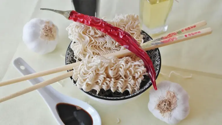 A bowl of curly ramen noodles with chopsticks, surrounded by garlic bulbs and a bright red chili pepper on a light surface.