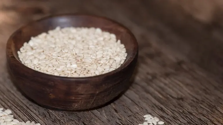 A wooden bowl filled with sesame seeds on a textured wooden surface