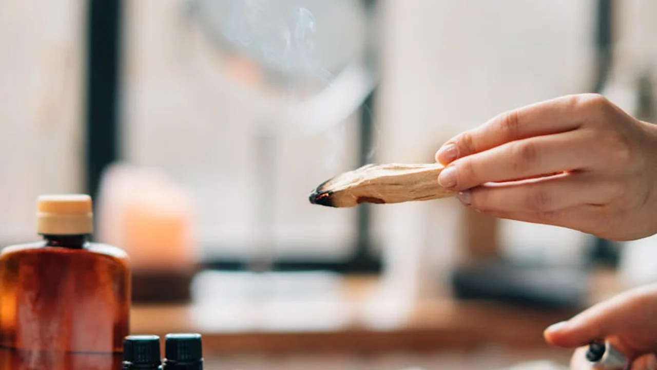A close-up of a hand holding a small wooden scoop near a bottle of essential oil, implying adding fragrance to sauna steam.