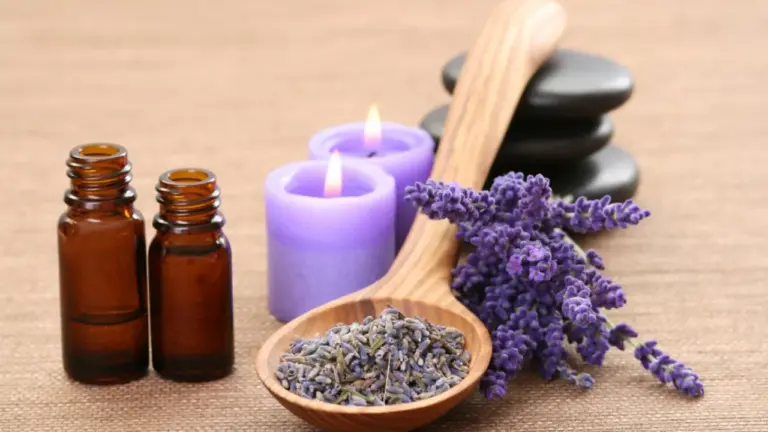 Brown glass essential oil bottles with a wooden spoon filled with dried lavender, lavender sprigs, and purple candles on a wooden surface.