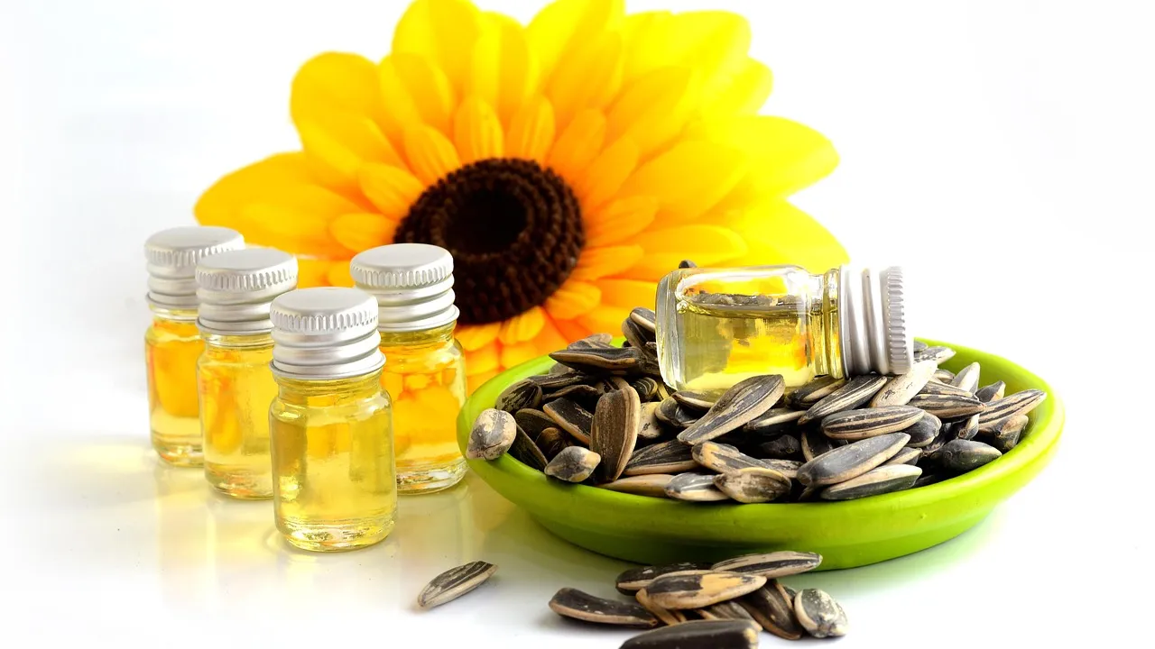 A bright sunflower in the background with small bottles of cooking oil and a green bowl filled with seeds on a white surface.