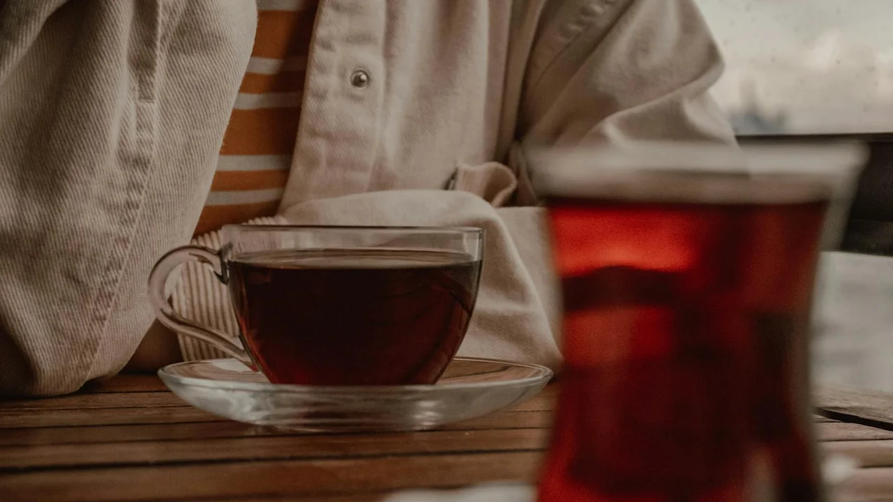 A person seated at a wooden table with a cup of tea on a saucer; a blurred glass of tea appears in the foreground.