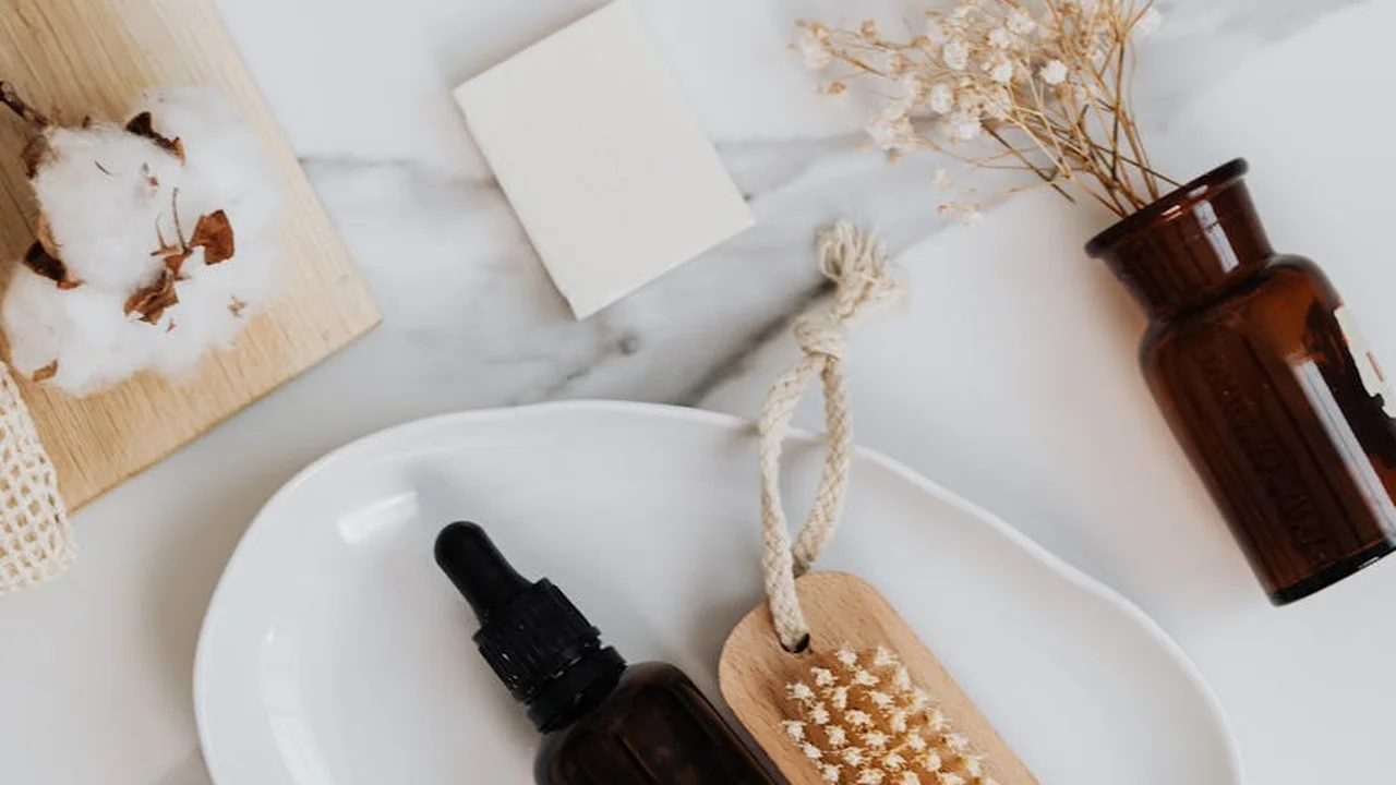 Flat lay of tea tree oil hair care setup on a white marble surface: a brown glass dropper bottle, a bar of soap, cotton, and dried flowers.