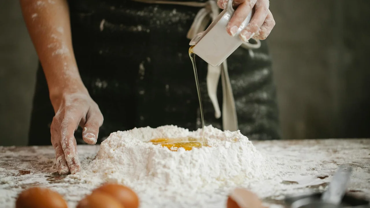 A cook wearing an apron sprinkles oil from a measuring jug onto a mound of flour on a cluttered kitchen counter.