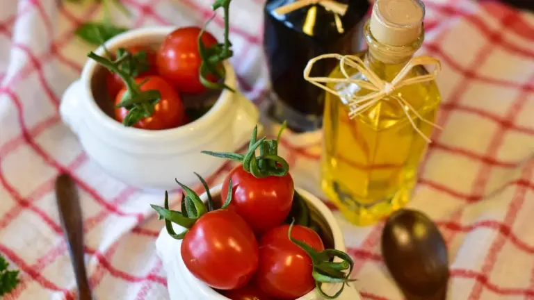 Tomatoes in white bowls with a bottle of oil on a checkered tablecloth.