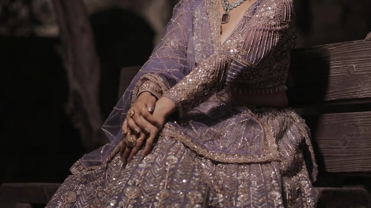 Individual seated on a wooden bench wearing an ornate purple dress and sparkling jewelry.
