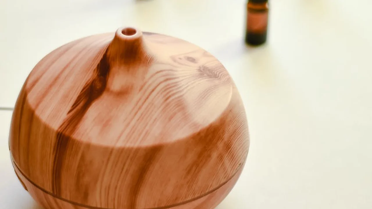 Wooden aroma diffuser with visible wood grain on a light surface; a small bottle appears blurred in the background.