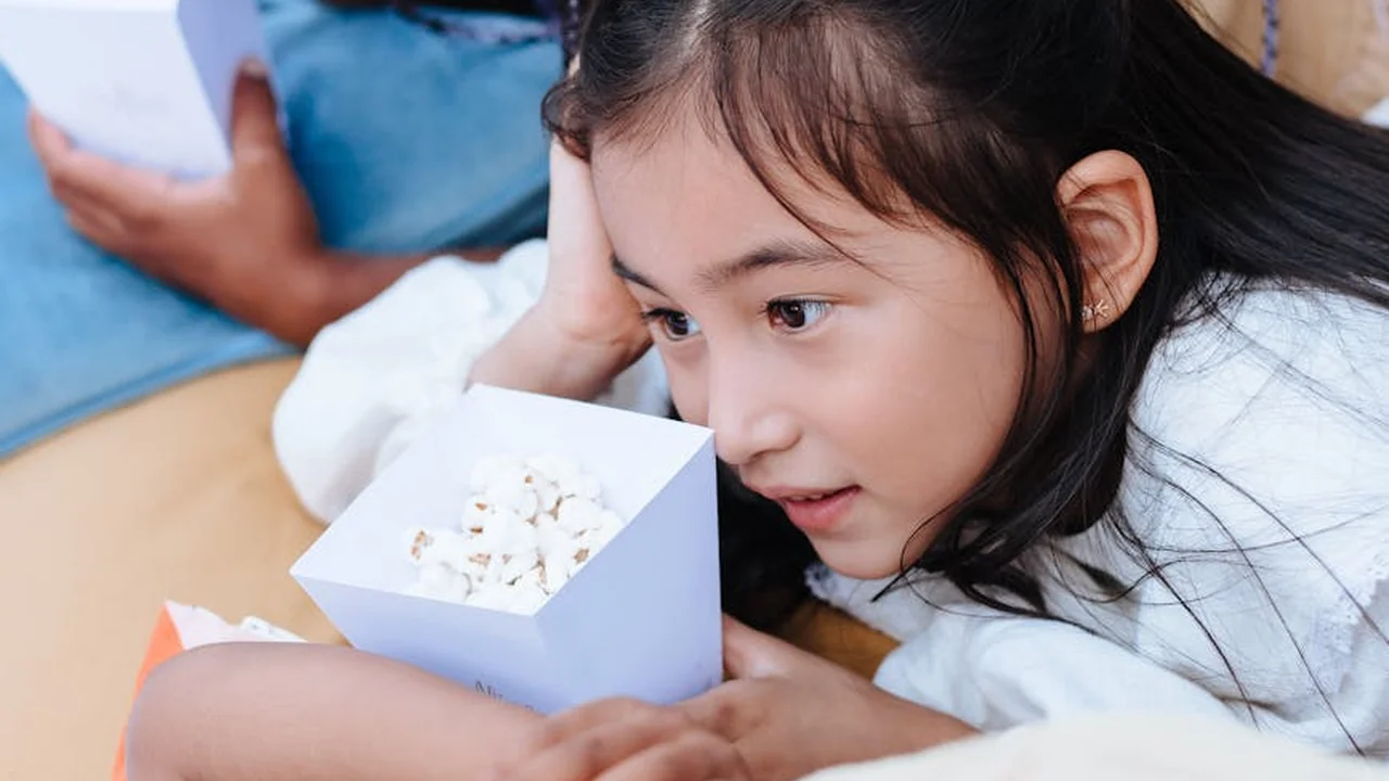 Close-up of a young girl holding a small box of popcorn while lying on a sofa, with another person blurred in the background.